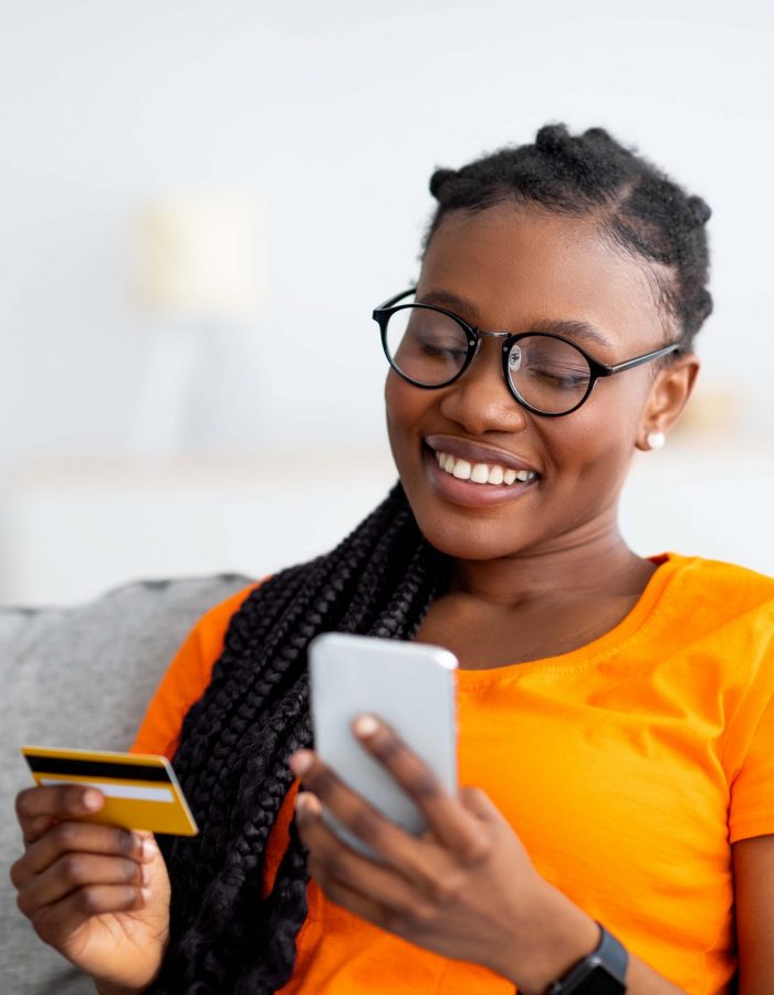 cheerful-young-black-woman-purchasing-on-web-from-home-sitting-on-sofa-using-smartphone-and-credit.jpg